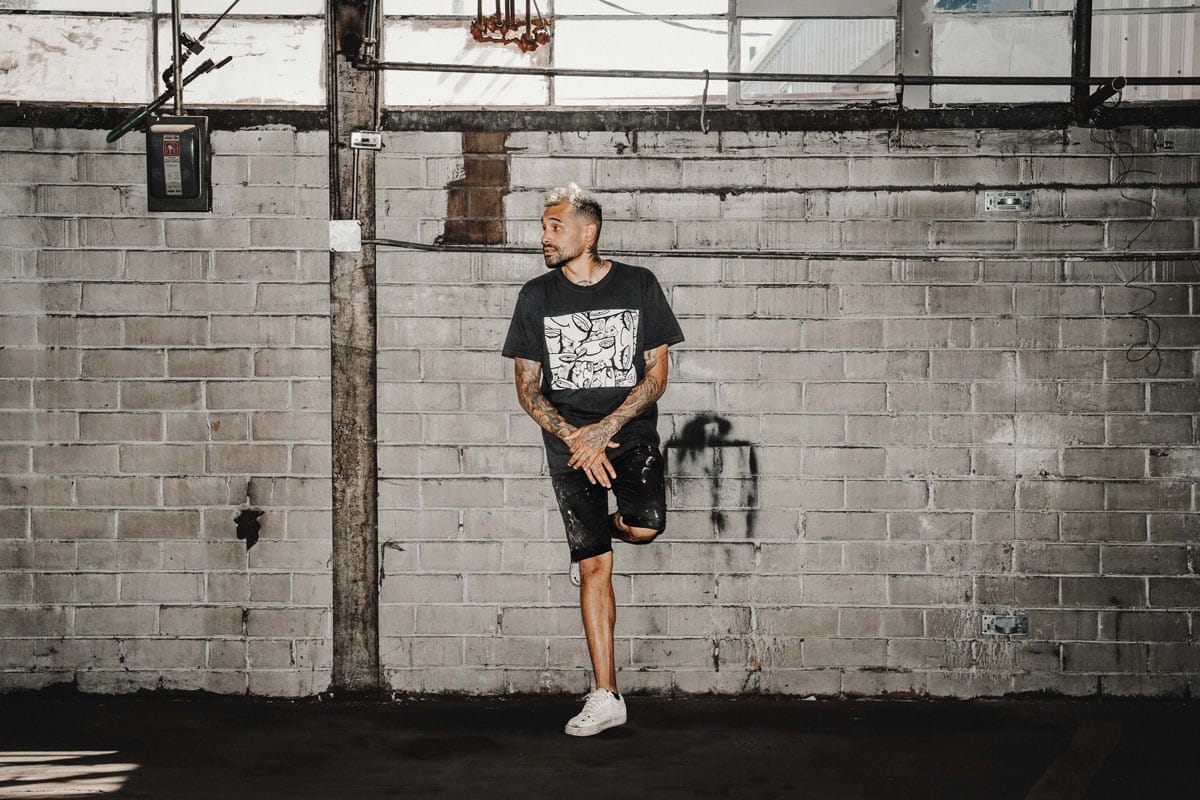 the artist RubeZilla poses in a black shirt standing against a brick wall